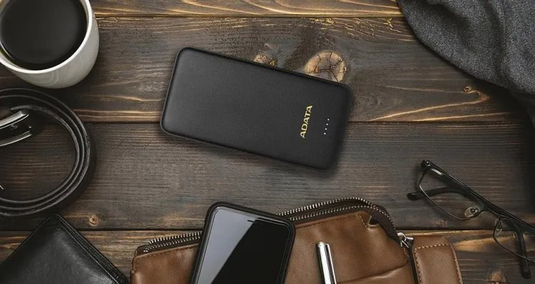 پاوربانک ADATA T10000 روی میز چوبی در کنار کیف، گوشی هوشمند، عینک و لیوان قهوه